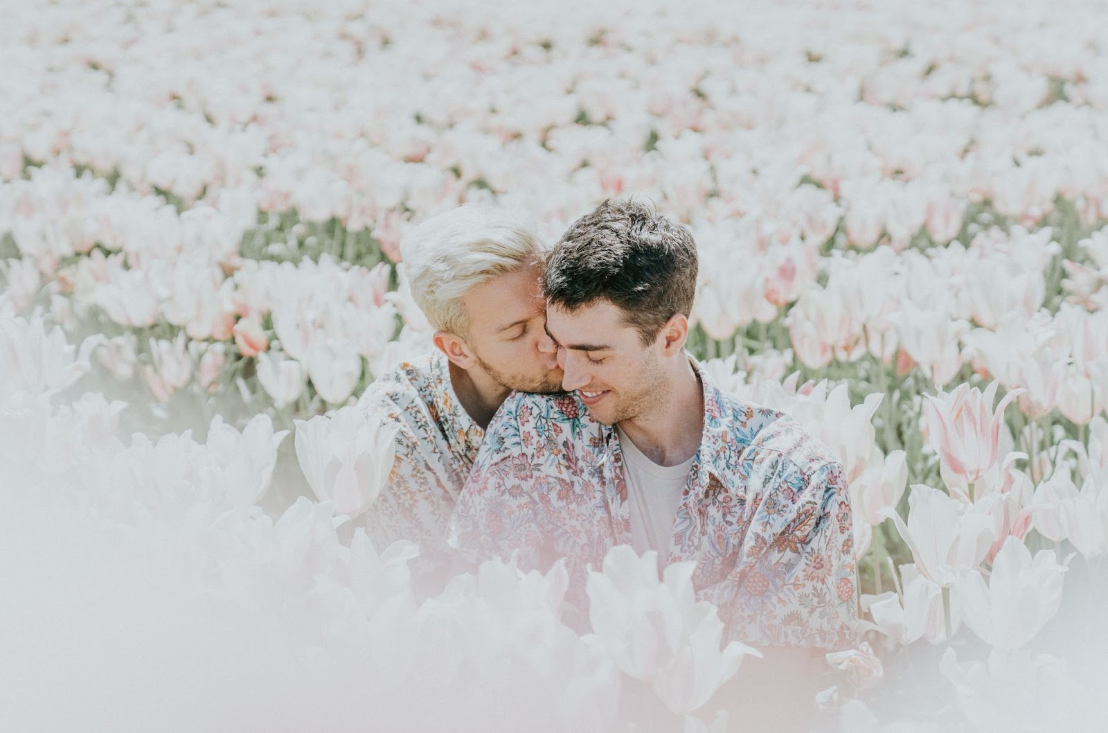 couple in matching engagement photoshoot outfits sitting in a field of flowers while one kisses the other's cheek
