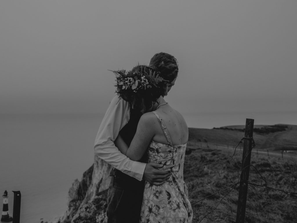 black and white photo of a couple hugging while standing on a cliff overlooking the water 
